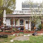 Imbiss Asia mit Holzmöbeln und bunten Blumen, Baum im Blütenstand, im Hintergrund ein modernes Gebäude.