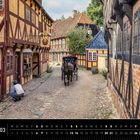 Dänemark 03. Eine historische Straße mit Fachwerkhäusern, Kopfsteinpflaster, Kutsche, Person und dekorativem Schild.