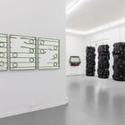 Drei grüne, lineare Kunstwerke an der Wand, daneben vier hängende schwarze Skulpturen und ein roter, ramponierter Autoteil.