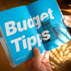 „Budget Tipps“ und „Kapitel 6“ stehen in großer, weißer Schrift auf blauem Hintergrund neben einem Münzstapel. Hände halten das Buch.