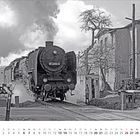 Kalendertext: März. Ein schwarzer Zug mit der Nummer 01 2204-4 auf einem Bahnübergang, Bäume und ein Gebäude im Hintergrund.