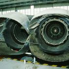 Zwei überdimensionierte, mechanische Düsentriebwerksauslässe in einem Hangar, umgeben von Werkzeugen und Bodenmarkierungen.