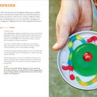 "GÖTTERSPEISE" steht oben. Ein Teller mit grüner Götterspeise, dekoriert mit bunten Gummiwürmern und einer roten Kirsche.