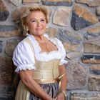 Eine ältere Frau in traditioneller bayerischer Tracht vor einer Natursteinwand. Sie lächelt freundlich.