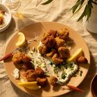 Frittierte Häppchen mit Zitronenscheiben und Dip auf einem Teller, umgeben von Pflanzen und einem Getränk.