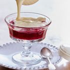 Ein Glas mit roter Fruchtcreme, auf das dicke, helle Sauce gegossen wird. Eine kleine Löffel liegt auf einem Teller daneben.