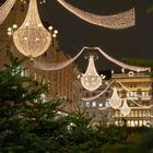 Lichterketten und große, leuchtende Kronleuchter in der Nacht über einer von Tannen gesäumten Einkaufsstraße.