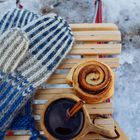 Gestrickte Fäustlinge neben einer Zimtschnecke und einer Tasse mit Zimt, auf einem Holzschlitten im Schnee.