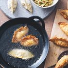 Gyoza in einer Pfanne werden frittiert. Daneben liegen ungegarte Gyoza und eine Schüssel mit gehacktem Gemüse.