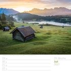 29. Woche, Juli 2027. Holzhütten in grüner Landschaft vor Bergen bei Sonnenaufgang. Kalenderseite mit Datum und Wochentagen.