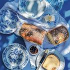 Drei blau-weiße Teller, Brot mit Körnern, Butterstückchen unter Glas, Marmelade, bestrichenes Brot mit Messer auf Tischdecke.