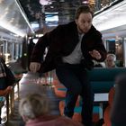 Kein sichtbarer Text. Eine Person springt in einem modernen Zugabteil, umgeben von sitzenden Passagieren.