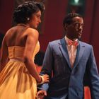 Ein Paar in eleganter Kleidung steht in einem Theater. Sie trägt ein gelbes Kleid; er einen blauen Anzug mit rosa Fliege.