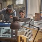 Ein Mann mit "BIG BUCK"-Mütze zeigt einer Frau etwas auf einem Monitor in einem Büro mit mehreren Bildschirmen.