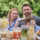 Ich kann keine Texte oder Namen erkennen. Zu sehen ist eine Familie draußen am Tisch mit Getränken, Obst und einer Blume.