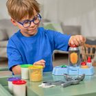 Ein Kind mit blauer Brille spielt an einem Tisch mit bunten Dosen und einem wissenschaftlichen Spielzeug-Set.