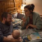 Ein Mann und eine Frau im Bett mit einem Baby. Die Frau gestikuliert, während ein Buch mit Text vor ihr liegt. Gemütliche Stimmung.