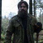 Es gibt keinen sichtbaren Text. Ein bärtiger Mann in einer Jacke steht in einem Wald und hält einen Stock.