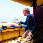 Ein Mann kniet in einem Rettungsboot und blickt aufs Meer; im Hintergrund ein sinkendes Schiff.