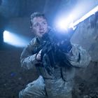 Ein Soldat in Tarnkleidung kniet mit einer Waffe und einem hellen Lichtschein in einer dunklen Höhle.