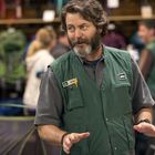 Ein Mann mit Bart trägt eine grüne Weste mit den Aufschriften "Dan" und "REI". Im Hintergrund sind Rucksäcke sichtbar.