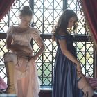 Zwei Frauen in historischen Kleidern stehen vor einem verzierten Fenster. Sie befestigen Strumpfhalter an ihren Beinen.