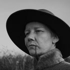 Eine Person mit ernstem Blick trägt einen breiten Hut und einen groben Mantel, im Hintergrund ist hohes Gras zu sehen.
