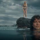 Zwei Frauen am Meer, eine schwimmt im Wasser, die andere steht auf einem Felsen. Der Himmel ist bewölkt.