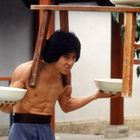 Eine muskulöse Person balanciert Schalen und einen Schemel, der auf den Schultern ruht, in traditioneller Umgebung.