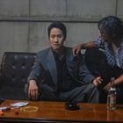 Zwei Männer sitzen in einem düsteren Raum; einer im Anzug mit Zigarette. Ein Tisch vor ihnen mit Flasche und kleinen Gegenständen.