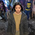 Ein Mädchen in einer dicken Jacke schaut besorgt, umgeben von Menschen in bunter Kleidung, die unklar im Hintergrund sind.