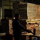 Ein Mann in schwarzer Lederjacke steht mit einer Waffe in einem Lager voller gestapelter Kartons.