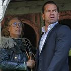 Zwei Männer stehen besorgt nebeneinander vor einem rustikalen Gebäude. Einer trägt eine Brille und ungewöhnliche Kleidung.