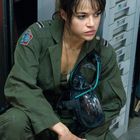 Eine Frau in grüner Uniform kauert. Aufgenähtes Emblem links, Helm in der Hand. Sie sieht nachdenklich aus.