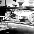 „Das Rätsel der roten Orchidee“ steht in einem Auto, ein Mann und eine Frau sitzen im Cabrio und schauen nach vorne.