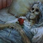Eine Skelettfigur mit langem grauem Haar liegt im Bett, hält eine rote Rose und trägt ein weißes Kleid.