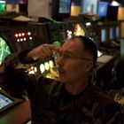 Ein Mann in Militäruniform zeigt auf einen Radarschirm in einem Kontrollraum mit grünen Karten und blinkenden Lichtern.