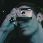 Person hält eine silberne Kamera vor dem Gesicht, dunkel beleuchtete Straße im Hintergrund.