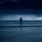 Ein einsamer Mensch steht am dunklen Strand, dem Ozean zugewandt, unter einem bedrohlichen, dunklen Himmel.
