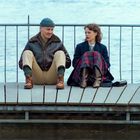 Ein Mann und eine Frau sitzen auf einem Steg am Wasser, lehnen sich an ein Geländer und tragen warme Kleidung.