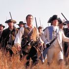 Eine Gruppe von Männern in historischen Uniformen marschiert durch hohes Gras, einige tragen Musketen und Hüte.