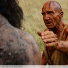 Ein Mann, rotbraun bemalt, streckt die Hand aus, vor ihm eine große, dunkle Kreatur, in einer Graslandschaft.