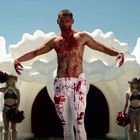 Ein blutverschmierter, muskulöser Mann vor weißer, wolkenförmiger Kulisse, flankiert von zwei Cheerleaderinnen.