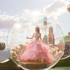 Keine erkennbaren Texte. Eine Frau in einem rosa Kleid steht in einer schimmernden Kugel vor einer fantasievollen, märchenhaften Kulisse.