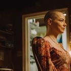 Eine Frau mit rasiertem Kopf trägt ein gemustertes Kleid in einem Raum voller Regale und Urkunden. Warmes Licht erhellt die Szene.