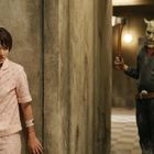 Kein Text enthalten. Eine Person in rosa Pyjama wirkt ängstlich, während eine maskierte Figur mit Axt in einem düsteren Flur steht.