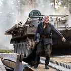 Ein Mann mit grauem Haar, zerlumpter Kleidung vor einem rauchenden Panzer im Wald, trägt eine Stiefelsohle.