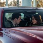 Zwei Personen im Auto, sie blicken sich ernst an. Der Wagen ist rot. Hintergrund: unscharf, mit Bäumen und Strukturen.