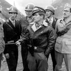 Ein Soldat in Lederjacke zeigt mit einem Stock. Sechs Männer tragen Uniformen und Helme, stehen in bergigem Gelände.