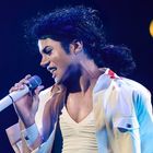 Person mit lockigem Haar singt leidenschaftlich in ein Mikrofon, trägt eine weiße Jacke und ein Licht strahlt auf sie.